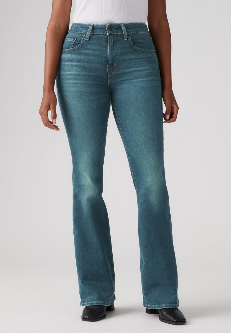 Jeans flare blu in denim con una finitura sbiadita, vita alta, tasche frontali e dettagli di cucitura in evidenza. Indossati con stivaletti neri alla caviglia.