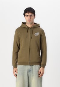 Olive zelena zip-up kapuca iz mehkega materiala, z logotipom spredaj, kapuco z vrvico in kenguru žepom. Standardna prileganje z rebrastimi manšetami.