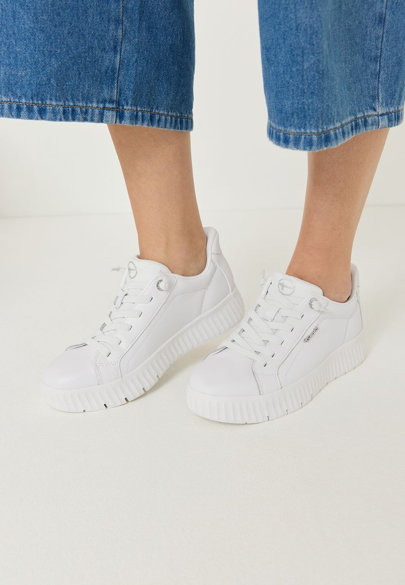 Άτομο που φοράει λευκά χαμηλά sneakers με χοντρές σόλες και κορδόνια, συνδυασμένα με μπλε κοντά τζιν σε λευκό πάτωμα.
