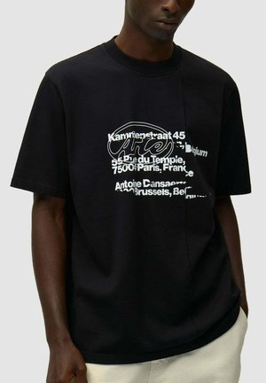 Uomo che indossa una t-shirt nera oversize con testo astratto bianco e indirizzi stampati sul davanti, abbinata a pantaloni chiari.