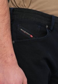 Ciemnoniebieskie jeansy z denimu z kontrastującym czarnym pasem. Posiadają przednią kieszeń z czerwoną i białą metką marki, akcentowaną niebieskim przeszyciem.