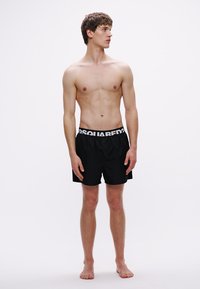 Bañador corto negro con cintura elástica marcada con la marca. Los shorts son lisos y tienen un diseño de corte holgado. El modelo está descalzo, mirando hacia la derecha.