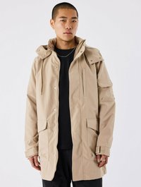 Beige hættetrøje med en tekstureret finish, der har trykknaplukninger, forlommer og justerbare snore. Båret over en sort shirt.
