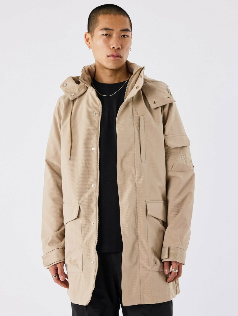 Beige hættetrøje med en tekstureret finish, der har trykknaplukninger, forlommer og justerbare snore. Båret over en sort shirt.