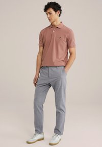 Polo de color rosa empolvado, de manga corta, con un logo negro. Combinado con unos pantalones claros de textura gris y zapatillas blancas con acentos beige.