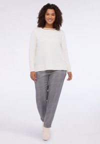 Weißer Strickpullover mit geripptem Saum und goldenen Akzenten am Kragen, kombiniert mit grauen Karohosen und beigen Knöchelstiefeln.