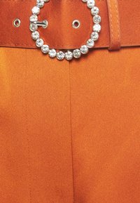 Oranje stoffen riem met een ronde gesp versierd met heldere edelstenen, met een gladde textuur en vlak ontwerp. Twee metalen verstelgaten zichtbaar.