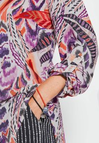Blouse colorée avec des motifs floraux violets, oranges et noirs ; tissu léger ; manches retroussées fixées par des boutons ; détail de bracelet superposé.