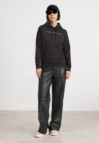 Sudadera negra con el logo "TOMMY JEANS", pantalones de cuero negro a juego y zapatillas ligeras. Accesorios incluyen una gorra negra y gafas de sol.