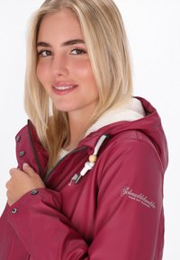 Bordo spalvota vandeniui atspari striukė su minkšta fliso pamušalu, įtvirtinimu su virvute ir logotipo detale ant rankovės; turi spaustukus ir užtrauktuką.