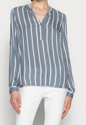 Mujer con blusa azul de rayas blancas verticales y pantalones blancos, de pie contra un fondo claro y liso.