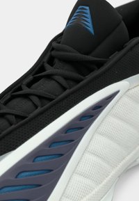Melns sporta apavs ar teksturētu materiālu, zilām detaļām, veidotu baltu sānu paneli un izsmalcinātu saķeres rakstu zolē.