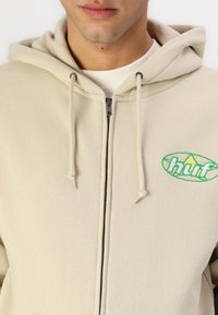 Bežová mikina s kapucňou a zipsom vpredu, s zeleným logom na boku. Materiál je štruktúrovaná tkanina s akcentmi šnúrky.