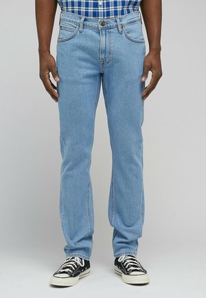Uomo con jeans azzurri a gamba dritta, sneakers nere e bianche e una camicia a quadri blu, in piedi davanti a uno sfondo semplice.