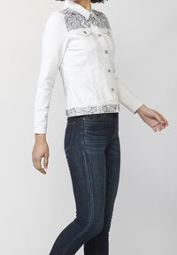 Biała kurtka jeansowa z wzorzystym czarno-białym kołnierzem i dolnym obszyciem, z zapięciem na guziki i dwoma kieszeniami na piersi. W połączeniu z ciemnoniebieskimi dżinsami.