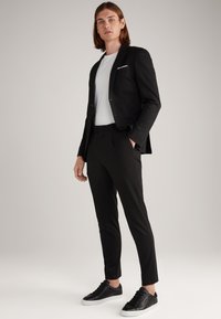 Schwarze maßgeschneiderte Sakko über einem weißen T-Shirt, kombiniert mit schwarzen Slim-Fit-Hosen und schwarzen Sneakers, die minimalistische Designelemente aufweisen.