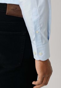 Blizu prikaz ruke osobe pored crnih traperica s smešnim kožnim oznakom Hackett London i manžetom svijetloplavog dugog rukava.