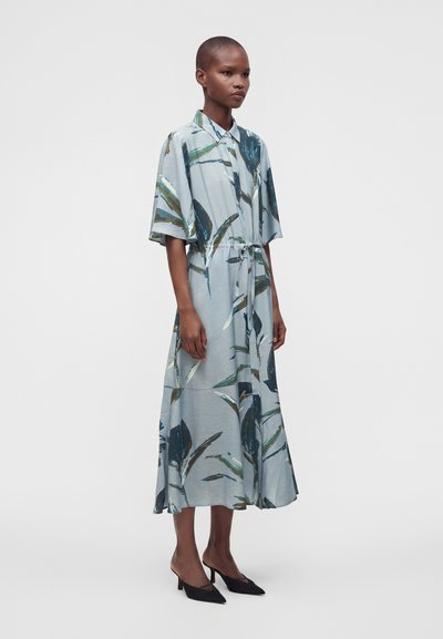Femme portant une robe midi bleu clair à imprimé botanique, manches courtes, devant boutonné, et des chaussures noires à talons bas et pointues.