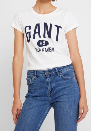 Mujer con una camiseta blanca de manga corta con el texto "GANT 49 New Haven" y vaqueros azules de tiro alto sobre un fondo liso.