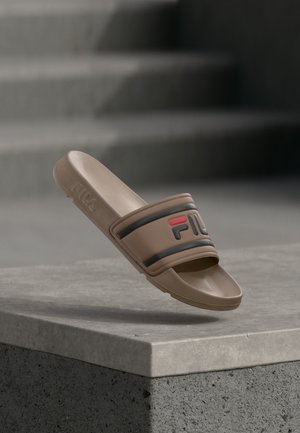Braune FILA Slide-Sandale mit schwarzem und rotem Logo, die am Rand einer grauen Betonstufe mit Treppen im Hintergrund liegt.