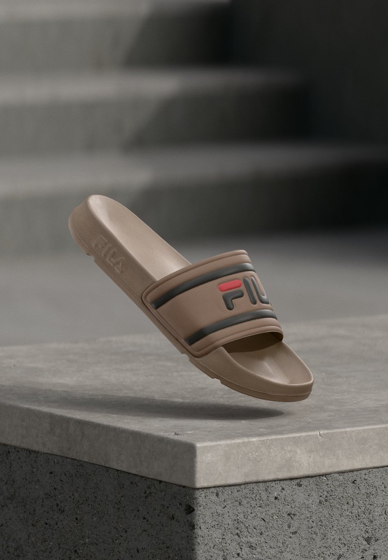 Καφέ σανδάλι FILA slide με μαύρο και κόκκινο λογότυπο, ακουμπισμένο στην άκρη ενός γκρι σκυρόδεματος σκαλοπατιού με σκάλες στο φόντο.