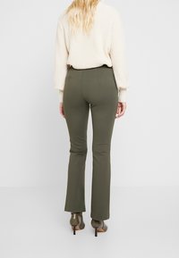 Olijfgroene, uitlopende broeken van rekbaar materiaal, gecombineerd met een beige gebreide trui, gezien van achteren.