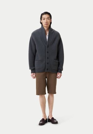 Cardigan gris charbon avec un col châle, fermeture par boutons et poches avant, associé à un short marron et des mocassins foncés.