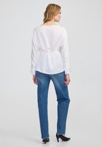 Vrouw gezien van achteren, gekleed in een witte blouse met lange mouwen die aan de achterkant is vastgebonden, blauwe spijkerbroek en zwarte hoge hakken, staand op een effen achtergrond.