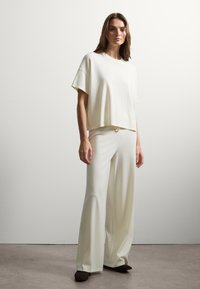 Set di loungewear color crema chiaro composto da una maglietta corta a maniche corte e pantaloni a gamba larga, realizzato in morbido tessuto testurizzato. Scarpe scure completano il look.