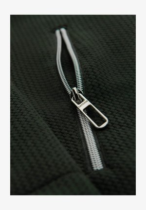 Tissu noir texturé avec fermeture éclair ; tirette de fermeture éclair en argent brillant avec marquage ; le tissu présente un motif de tissage.
