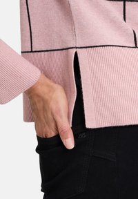 Helles rosa Sweatshirt mit schwarzen geometrischen Linien, gerippten Bündchen und seitlichen Schlitzen. Schwarze Hosen mit sichtbarem Taschendesign und Logo-Detail.