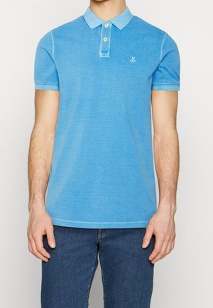 Mand iført en lyseblå poloshirt med korte ærmer og to knapper samt mørkeblå jeans, stående foran en ensfarvet baggrund.