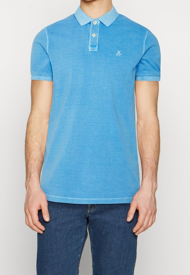 Homme portant un polo bleu clair à manches courtes avec deux boutons et un jean bleu foncé, debout devant un fond uni.