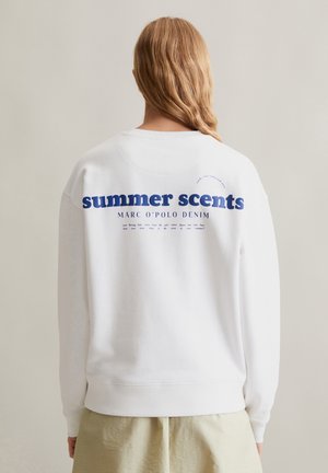 Person mit langem blondem Haar trägt weißen Pullover mit blauem Schriftzug „summer scents“ und beige Hose, vor einfarbig hellem Hintergrund.