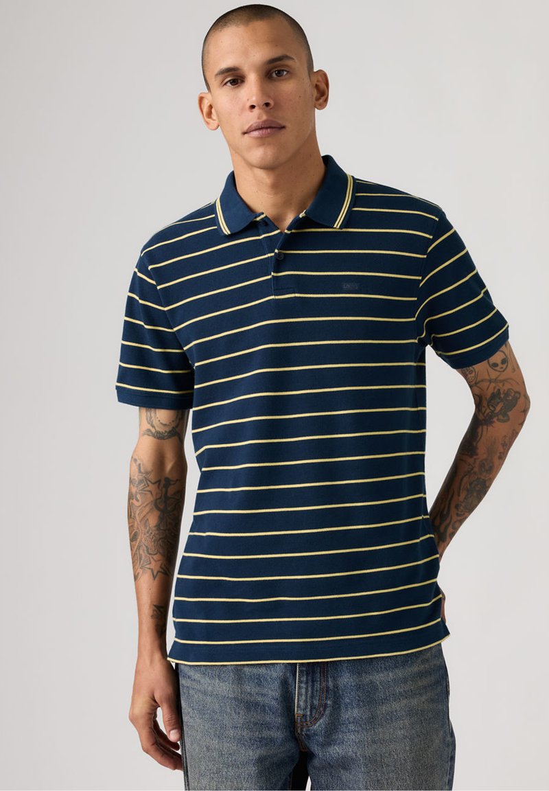 Mand med tatoveringer iført en marineblå polo med tynde gule vandrette striber og blå jeans, stående med den ene hånd bag ryggen.