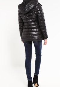Chaqueta acolchada negra con capucha, acabado brillante, diseño acolchado y puños elásticos, llevada sobre jeans oscuros y botas negras hasta el tobillo.