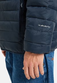 Detailný záber na ruku osoby blízko manžety námorníckej modrej prešívanej bundy Quiksilver a vrecka modrých džínov.