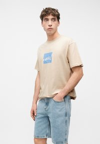 Νέος άντρας που φοράει μπεζ μπλουζάκι με σχέδιο και ανοιχτόχρωμα τζιν σορτς, στέκεται με το ένα χέρι στην τσέπη, κοιτώντας προς τα πλάγια μπροστά σε απλό φόντο.