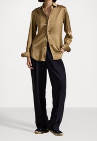 Gul satin långärmad skjorta med knäppning framtill och manschetter, i kombination med löst sittande svarta byxor och marinblå espadrillos.