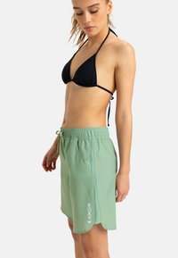 Zelená plavková sukňa s elastickým pásom a bočnými vreckami, s členitým lemom a bielym logom "ROXY" na prednej strane. Kombinovaná s čiernym bikiny topom.