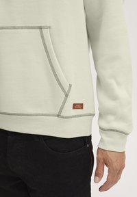 Sudadera verde claro con un bolsillo delantero, costuras en contraste y una pequeña etiqueta marrón en el lado inferior; combinada con pantalones negros.