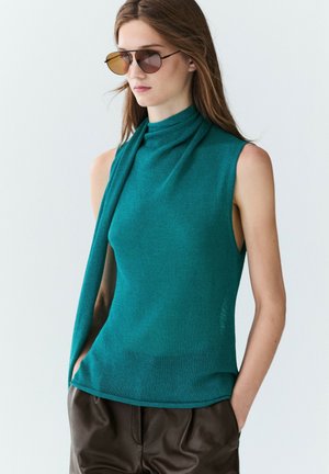 Donna con occhiali da sole, top smanicato color verde petrolio con dettaglio a sciarpa e pantaloni scuri, in piedi su uno sfondo chiaro e semplice.