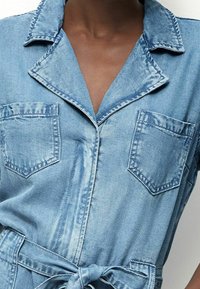Denim overal s světle modrým omytem, strukturovaným límcem, dvěma náprsními kapsami a páskem v pase. Měkká textura s volným střihem.