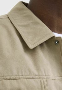 Helle beige Jacke aus einem glatten, strukturierten Stoff. Verfügt über einen spitzen Kragen, einen Druckknopfverschluss und sichtbare Nähte.