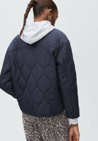 Mörkblå quiltad jacka med en vit hoodie under, med rundad halsringning och detaljer med tvåknappsärmar. Leopardmönstrade byxor syns nedanför.