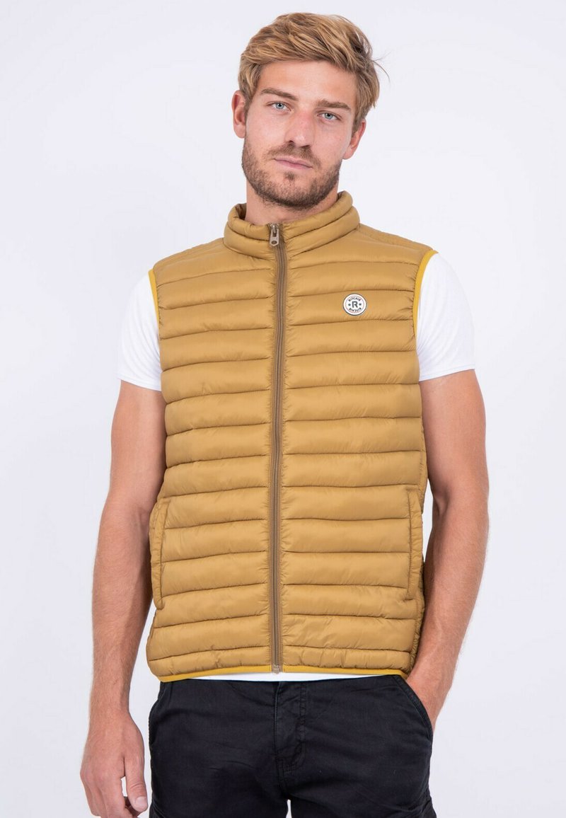 Gilet matelassé beige avec un col montant, un design à quilting vertical, une fermeture éclair à l'avant et un patch logo circulaire sur la poitrine gauche. Présente des accents jaunes.