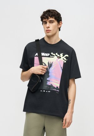 Młody mężczyzna z kręconymi włosami w czarnej koszulce Nike z grafiką, beżowych spodniach i czarnej torbie na ramię na jednolitym szarym tle.