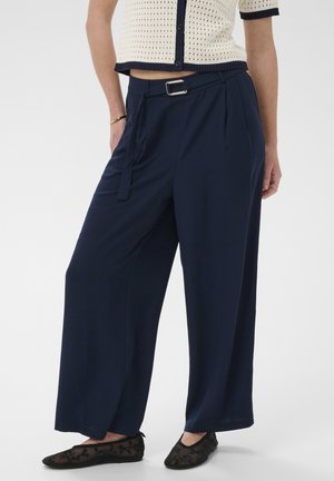 Nő sötétkék, széles szárú nadrágban övvel, krémszínű rövid ujjú kötött felsőben sötétkék szegéllyel, és fekete, virágmintás balerinacipőben.