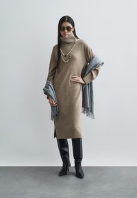 Bež pletena haljina s visokim rolkama, rebrastim rubom i bočnim prorezima. Uparena s crnim čizmama do koljena i sivim šal s resama.