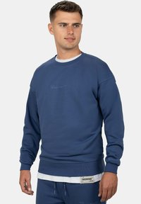 Felpa blu navy in un morbido tessuto con polsini e orlo a coste, con logo ricamato sul petto e una camicia bianca a strati sotto.